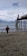 A child in a winter jacket and hat walks along a pebbly beach. A wooden pier extends over the water with an Argentine flag waving at its end. The background reveals a cloudy sky and distant mountains across the water.