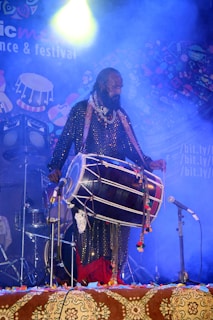 A musician stands on stage amidst colorful decorations and a smoky atmosphere, playing a large traditional drum. There are other musical instruments and stage lights in the background, and the setting suggests a vibrant festival or concert.