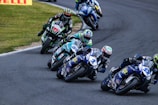 Several motorcycle racers lean into a curve on a paved racetrack. The riders are wearing colorful helmets and racing suits, while their motorcycles display various sponsor logos. The track is bordered by grass on one side with a few visible advertisements.