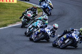 Several motorcycle racers lean into a curve on a paved racetrack. The riders are wearing colorful helmets and racing suits, while their motorcycles display various sponsor logos. The track is bordered by grass on one side with a few visible advertisements.