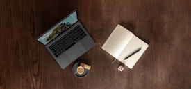 A wooden table surface with a sleek, open laptop displaying a nature-themed wallpaper. To the right of the laptop is an open notebook with a pen resting on it. A cup of coffee with a biscuit placed on the saucer and a small dice are also on the table.