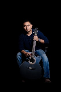 A person is seated with a guitar, wearing a dark shirt and jeans, illuminated against a black background. The individual holds the guitar in a relaxed manner, exuding a calm and focused demeanor. The lighting is dramatic, highlighting the person's facial features and the contours of the guitar.