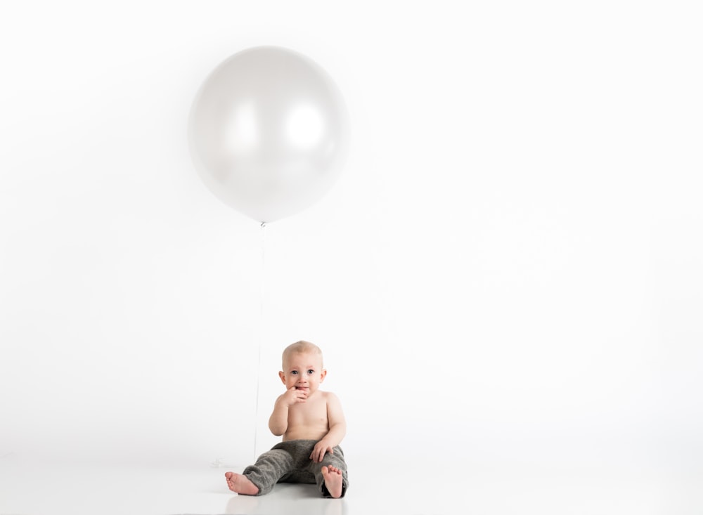 toddler in grey pants with white balloon sitting on floor