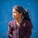 A young girl with long dark hair tied in a ponytail, wearing a knitted maroon sweater with a flower decoration, stands against a bright blue background. She looks to the side with a calm and contemplative expression.