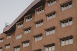 A multi-story building facade with several evenly spaced windows, featuring awnings above each window. The building has a light brown color with a slightly textured surface. The image captures the upper part of the building against a pale sky.