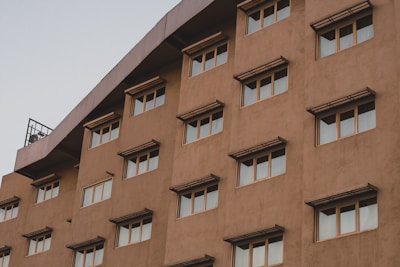 A multi-story building facade with several evenly spaced windows, featuring awnings above each window. The building has a light brown color with a slightly textured surface. The image captures the upper part of the building against a pale sky.