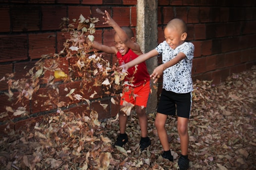 Two children play joyfully in a pile of dry leaves, with one wearing a red top and orange shorts and the other in a patterned shirt and black shorts. A brick wall provides the background, and the ground is covered with leaves.