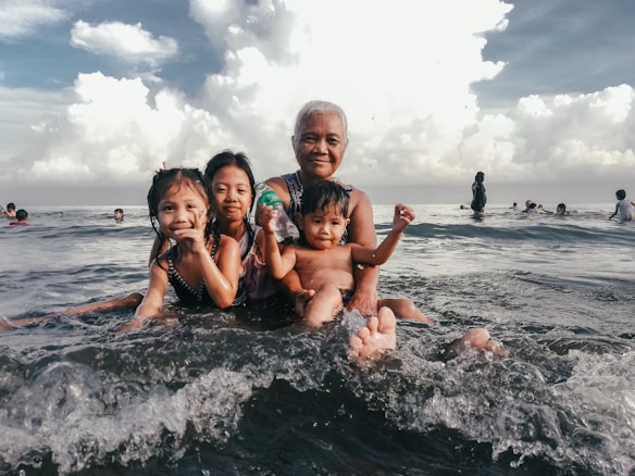 An elderly woman is in the water at the beach with three young children. They are all smiling and appear to be enjoying their time in the water. Other people can be seen in the background engaging in similar activities. The scene is calm with cloudy skies above.
