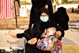 A woman wearing a face mask and black clothing is holding a sleeping child on her lap. The child is wrapped in a colorful blanket with vibrant designs. The setting appears to be outdoors with a few people in the background, some wearing similar black clothing.