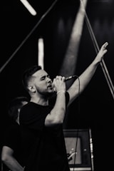 A male performer singing passionately into a microphone with one hand raised in the air, on a stage under dramatic lighting.