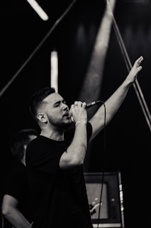 A male performer singing passionately into a microphone with one hand raised in the air, on a stage under dramatic lighting.