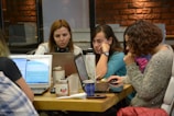 A group of women is sitting at a table, each using a laptop. Their expressions suggest focus and engagement. In the center, there are various items, including cups and a thermos. The setting appears to be a casual workspace with brick and glass walls in the background.