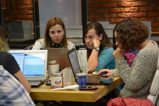 A group of women is sitting at a table, each using a laptop. Their expressions suggest focus and engagement. In the center, there are various items, including cups and a thermos. The setting appears to be a casual workspace with brick and glass walls in the background.