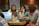 A group of women is sitting at a table, each using a laptop. Their expressions suggest focus and engagement. In the center, there are various items, including cups and a thermos. The setting appears to be a casual workspace with brick and glass walls in the background.