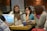 A group of women is sitting at a table, each using a laptop. Their expressions suggest focus and engagement. In the center, there are various items, including cups and a thermos. The setting appears to be a casual workspace with brick and glass walls in the background.