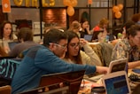 People are gathered in a busy workspace, collaborating and working on laptops. The room is filled with a diverse group of individuals, who appear focused and engaged in various activities. Orange balloons and decorations are visible, suggesting a festive or themed event.