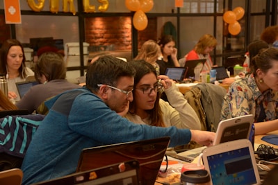 People are gathered in a busy workspace, collaborating and working on laptops. The room is filled with a diverse group of individuals, who appear focused and engaged in various activities. Orange balloons and decorations are visible, suggesting a festive or themed event.