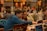 People are gathered in a busy workspace, collaborating and working on laptops. The room is filled with a diverse group of individuals, who appear focused and engaged in various activities. Orange balloons and decorations are visible, suggesting a festive or themed event.