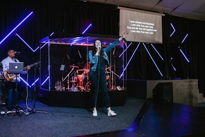 A band is performing on stage with a singer holding a microphone and raising one arm. The setting includes a guitarist on the left and a drummer positioned within a transparent enclosure. There are neon blue lights and a screen displaying song lyrics on the right.