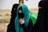 A group of women dressed in black garments with their faces covered, standing in an outdoor setting. One woman wears a turquoise hijab, adding a vibrant splash of color. They appear to be in a desert-like landscape, with muted and earthy tones in the background.