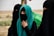 A group of women dressed in black garments with their faces covered, standing in an outdoor setting. One woman wears a turquoise hijab, adding a vibrant splash of color. They appear to be in a desert-like landscape, with muted and earthy tones in the background.
