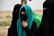 A group of women dressed in black garments with their faces covered, standing in an outdoor setting. One woman wears a turquoise hijab, adding a vibrant splash of color. They appear to be in a desert-like landscape, with muted and earthy tones in the background.