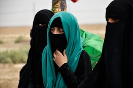 A group of women dressed in black garments with their faces covered, standing in an outdoor setting. One woman wears a turquoise hijab, adding a vibrant splash of color. They appear to be in a desert-like landscape, with muted and earthy tones in the background.