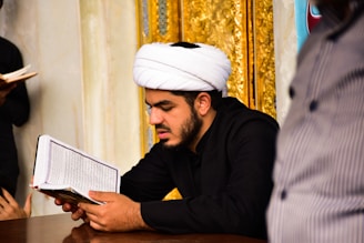A man wearing a traditional white turban and black clothing is reading a book with Arabic script. The background features ornate gold decorations.