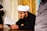 A man wearing a traditional white turban and black clothing is reading a book with Arabic script. The background features ornate gold decorations.