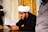 A man wearing a traditional white turban and black clothing is reading a book with Arabic script. The background features ornate gold decorations.
