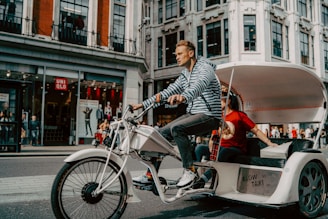 A person is pedaling a modern tricycle designed as a rickshaw or bike taxi along a city street. The tricycle has a striped canopy and white frame, with a passenger sitting behind the driver. The background features a storefront with mannequins and red signage, attracting pedestrians.