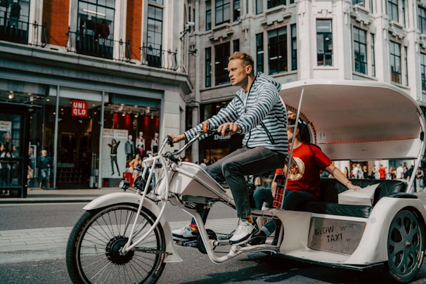A person is pedaling a modern tricycle designed as a rickshaw or bike taxi along a city street. The tricycle has a striped canopy and white frame, with a passenger sitting behind the driver. The background features a storefront with mannequins and red signage, attracting pedestrians.