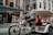A person is pedaling a modern tricycle designed as a rickshaw or bike taxi along a city street. The tricycle has a striped canopy and white frame, with a passenger sitting behind the driver. The background features a storefront with mannequins and red signage, attracting pedestrians.