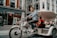 A person is pedaling a modern tricycle designed as a rickshaw or bike taxi along a city street. The tricycle has a striped canopy and white frame, with a passenger sitting behind the driver. The background features a storefront with mannequins and red signage, attracting pedestrians.
