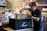 A person wearing a branded cap and shirt is placing a container into an insulated delivery bag. The scene appears to be inside a restaurant with a chef visible in the background near the kitchen. The environment is well-lit with hanging lights and has a modern ambiance.