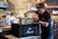 A person wearing a branded cap and shirt is placing a container into an insulated delivery bag. The scene appears to be inside a restaurant with a chef visible in the background near the kitchen. The environment is well-lit with hanging lights and has a modern ambiance.