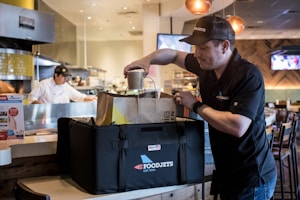 A person wearing a branded cap and shirt is placing a container into an insulated delivery bag. The scene appears to be inside a restaurant with a chef visible in the background near the kitchen. The environment is well-lit with hanging lights and has a modern ambiance.