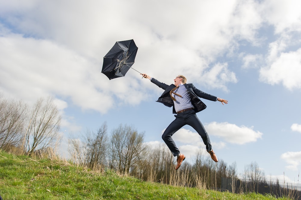 파란색과 흰색 하늘 아래 녹색 들판에서 점프하면서 우산을 들고 있는 남자