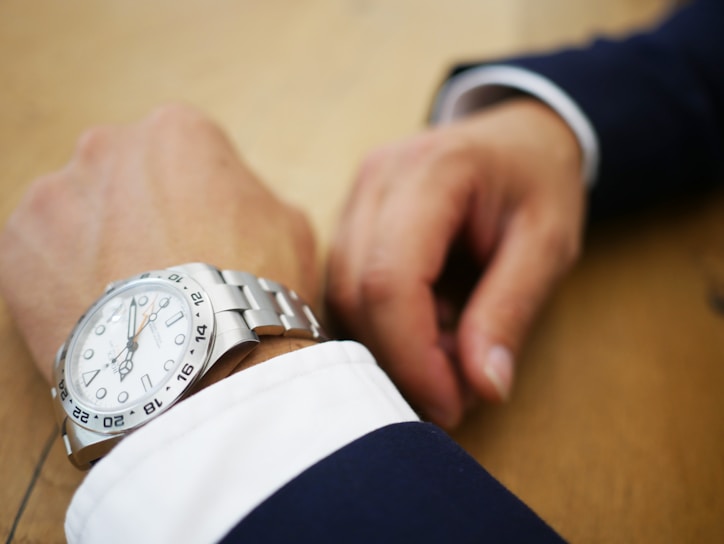 A close-up of a person's wrist adorned with a luxury silver wristwatch featuring a round face with hour markers and an orange second hand, set against a wooden surface. The person is wearing a dark suit with a white cuff, and the other hand is positioned nearby.