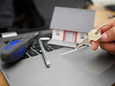 A model house sits on a laptop keyboard. Next to it, a screwdriver and an Allen key are placed, while a hand holds a set of keys, suggesting themes of homeownership and technology.