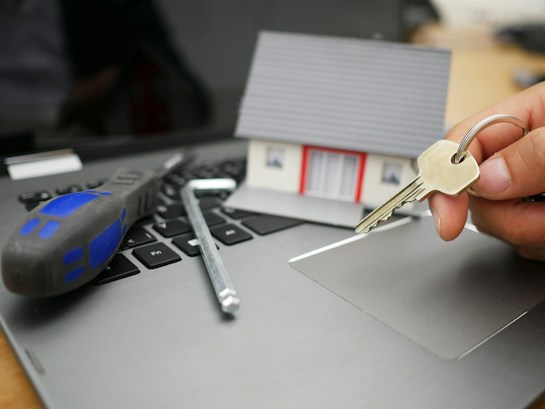 A model house sits on a laptop keyboard. Next to it, a screwdriver and an Allen key are placed, while a hand holds a set of keys, suggesting themes of homeownership and technology.