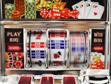 A close-up view of a slot machine with vibrant colors and various gambling symbols such as dice, coins, and playing cards. The reels display a combination of symbols, including bars, cherries, and the number seven. The chrome exterior and colorful details convey the theme of casino and gambling.