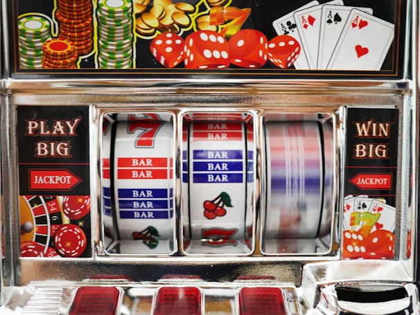 A close-up view of a slot machine with vibrant colors and various gambling symbols such as dice, coins, and playing cards. The reels display a combination of symbols, including bars, cherries, and the number seven. The chrome exterior and colorful details convey the theme of casino and gambling.