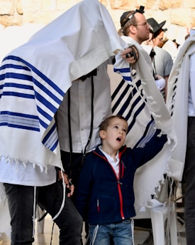 A group of people engage in a religious ceremony, with a young boy looking upward, standing partially under a prayer shawl held by someone. The atmosphere is busy with multiple participants wearing traditional attire, and some men have phylacteries on their heads. The setting appears to be a holy site.