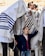 A group of people engage in a religious ceremony, with a young boy looking upward, standing partially under a prayer shawl held by someone. The atmosphere is busy with multiple participants wearing traditional attire, and some men have phylacteries on their heads. The setting appears to be a holy site.