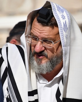 An elderly man with a gray beard and glasses is wearing traditional religious garments including a white cloth with black stripes and a head covering with a small black box on his forehead, known as a tefillin. Another person is partially visible in the background.