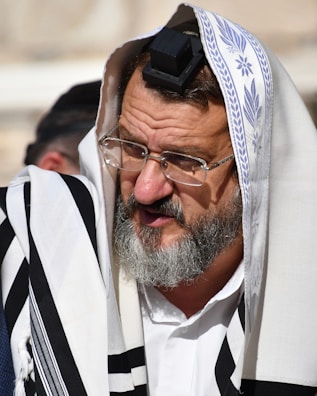 An elderly man with a gray beard and glasses is wearing traditional religious garments including a white cloth with black stripes and a head covering with a small black box on his forehead, known as a tefillin. Another person is partially visible in the background.