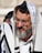 An elderly man with a gray beard and glasses is wearing traditional religious garments including a white cloth with black stripes and a head covering with a small black box on his forehead, known as a tefillin. Another person is partially visible in the background.