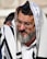 An elderly man with a gray beard and glasses is wearing traditional religious garments including a white cloth with black stripes and a head covering with a small black box on his forehead, known as a tefillin. Another person is partially visible in the background.