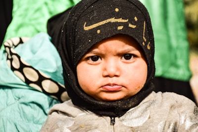 A young child with a thoughtful and serious expression, wearing a black headscarf with Arabic script. The child's jacket is gray with a shiny texture, and there is a background of green and black fabric.